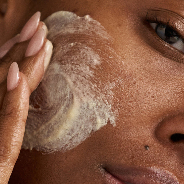 Close-up of a person applying a thick white cream or balm to their cheek with a finger, showing detailed skin texture and manicured nails.