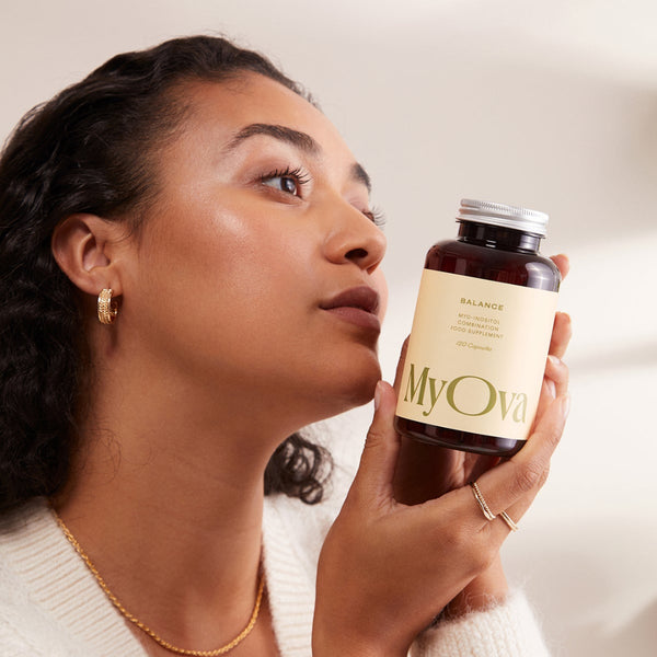 A woman in a cream knit cardigan holds a dark amber bottle labeled MyOva Balance, looking thoughtfully to the side against a soft beige background.