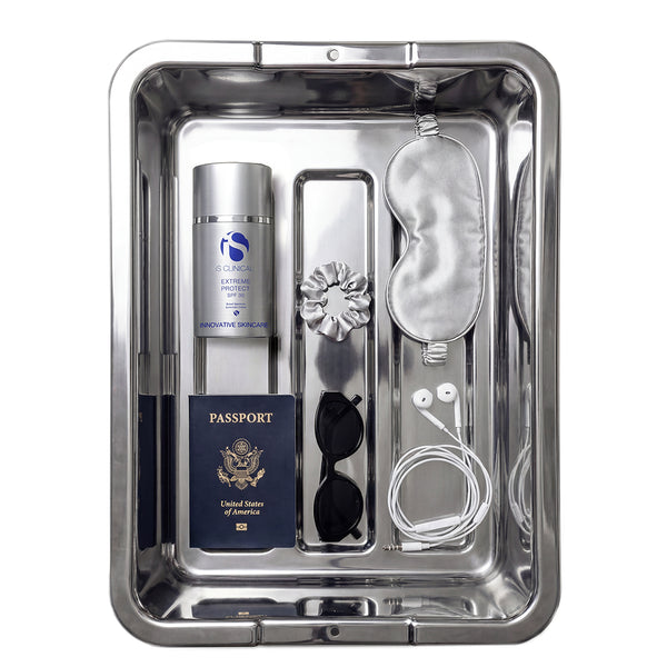 A stainless steel travel organizer tray containing a silver travel spray bottle, a navy blue United States passport, black sunglasses, white wired earbuds, a silver sleep mask, a silver flower-shaped hair clip, and a white cord, all arranged neatly on a white background.