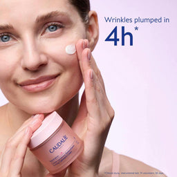 A woman applies cream to her cheek while holding a jar of Caudalie Deep Moisture Cream. The image features a light pink background with promotional text that reads 'Wrinkles plumped in 4h *'.