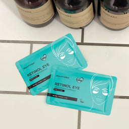 Two turquoise eye masks labeled 'Retinol Eye' on a tiled surface with bottles in the background.