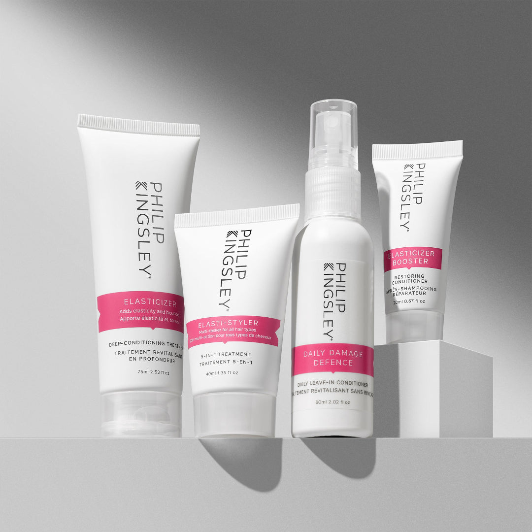 Four Philip Kingsley hair treatment products arranged on a light gray surface against a gray gradient background: an elasticizer treatment tube, an elasticizer styler tube, a daily damage defence spray bottle, and an elasticizer booster tube.
