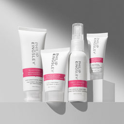 Four Philip Kingsley hair treatment products arranged on a light gray surface against a gray gradient background: an elasticizer treatment tube, an elasticizer styler tube, a daily damage defence spray bottle, and an elasticizer booster tube.