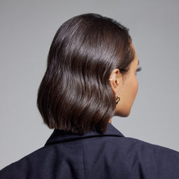 A woman shown from the back and side, wearing a dark navy blazer and a short, wavy dark brown bob haircut. She is wearing small gold hoop earrings against a solid gray studio background.