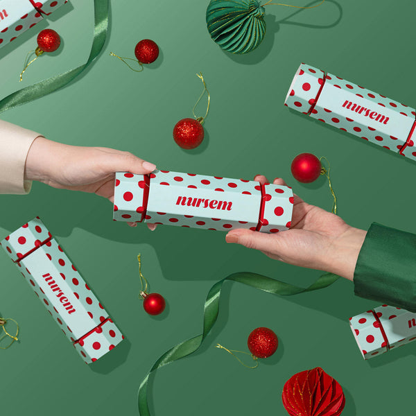 Two hands holding a white cylindrical Christmas nutcracker box with red polka dots and the brand name 'nurem' printed on it, set against a green background decorated with red ornaments, ribbons, and additional matching boxes.