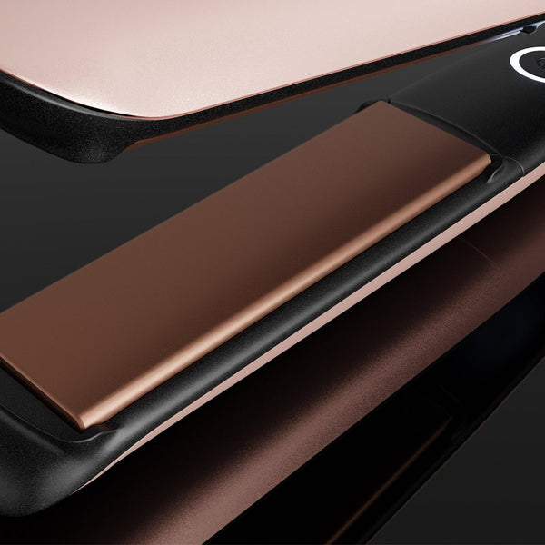Close-up of a hair straightener with metallic rose gold and black surfaces, shown on a dark reflective background.