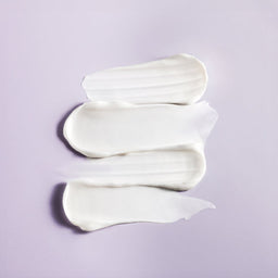 Three swatches of thick white cream or lotion arranged vertically on a light lavender background.