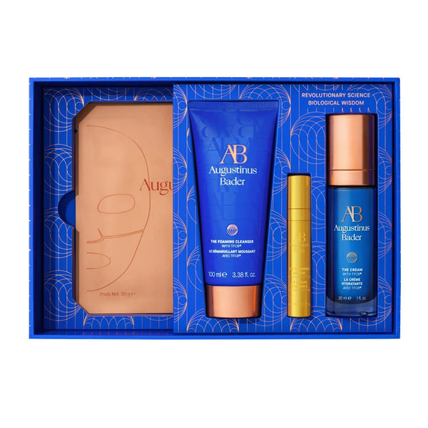 Augustinus Bader gift set in a blue box, featuring a tan leather-like item, a blue cleanser tube, a small gold tube, and a blue bottle with a rose-gold cap.