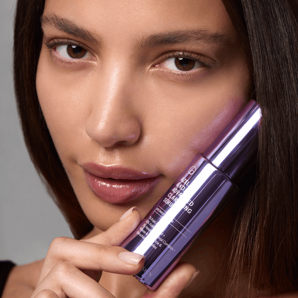 A close-up of a woman holding a metallic purple cosmetic tube against her cheek. The tube has white text that reads 'SKIN PERFECTING SERUM' and '10% PEPTIDE'.