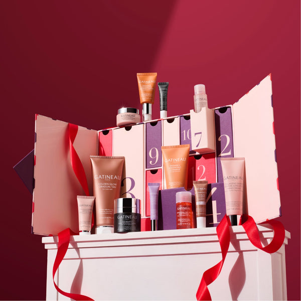 A Catineal skincare advent calendar set displayed on a white pedestal against a deep burgundy background. The set includes a large pink and white advent calendar box with numbered doors, several individual skincare products in pink, orange, purple, and black packaging, and red ribbon accents.