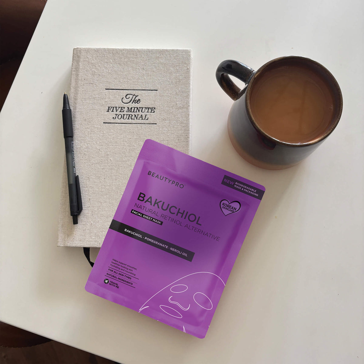 Purple skincare product package, notebook, pen, and mug on a white surface