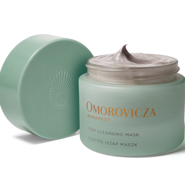 A jar of Omorovicza Deep Cleansing Mask with its lid off, showing the pale mint green jar filled with a thick, light-colored cream, and a matching lid leaning against it on a white background.