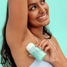 A woman holding a mint green and white deodorant stick under her arm against a solid light turquoise background.