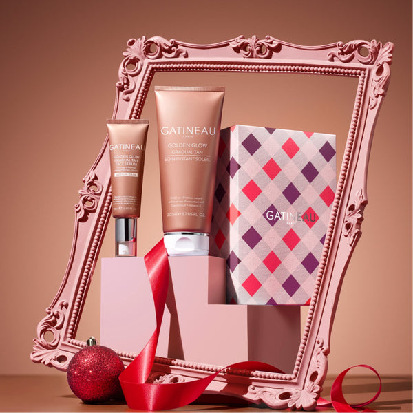 Gatineau Golden Glow body care set displayed in front of an ornate pink picture frame, featuring a body wash tube, a moisturizer tube, and a patterned box with a red ribbon and red ornament in the foreground.