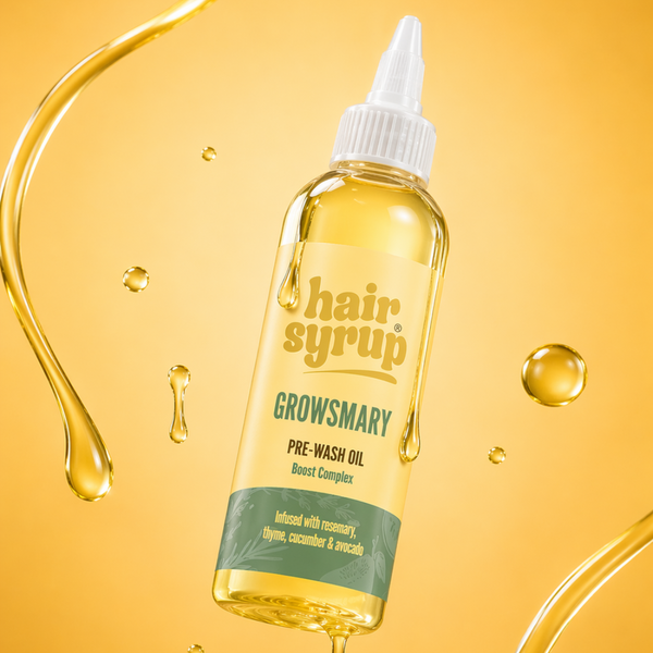 A bottle of Growsmary Hair Syrup pre-wash oil floating against a warm golden-yellow background with stylized liquid droplets and swirls. The bottle is clear, showing the golden-yellow oil inside, with a white dropper cap and a label featuring yellow, green, and white sections.