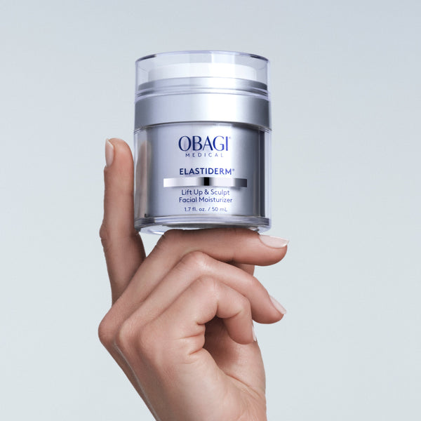 Hand holding a jar of Obagi Elastiderm cream against a light gray background