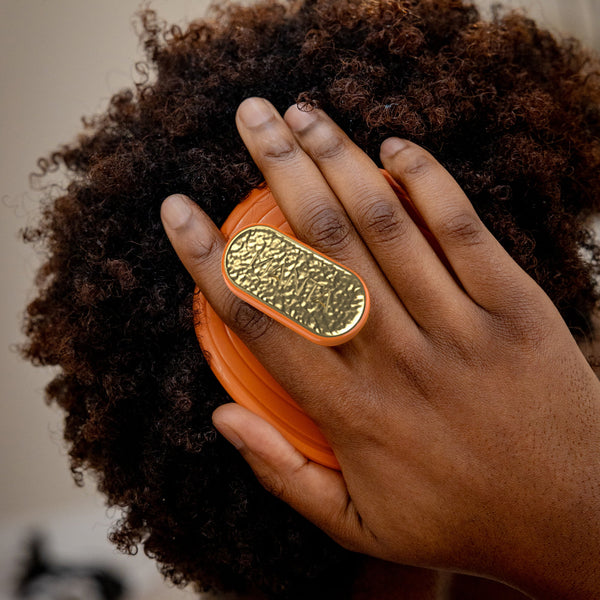 A close-up lifestyle shot of a person with dark curly hair wearing a large, chunky gold ring with an orange band. The ring features a textured, hammered oval face with a polished gold center.