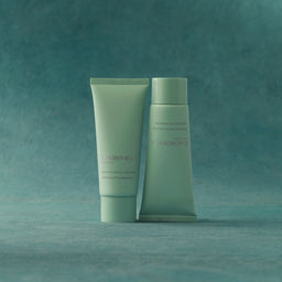 Two pale mint green cosmetic tubes from the brand Scionohydra, standing side-by-side on a textured teal surface against a matching teal background.