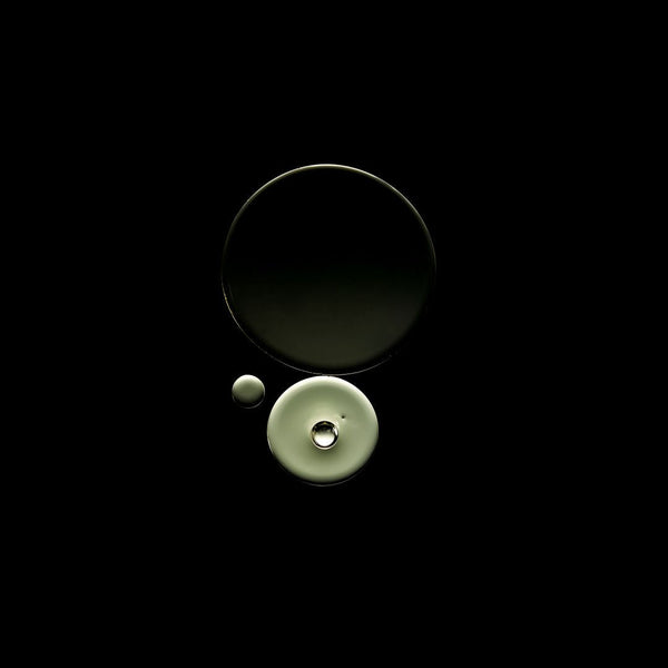 Three circular glass objects, including two larger dark-toned pieces and one pale greenish-beige piece with a small clear droplet, arranged on a solid black background.
