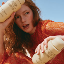 Woman holding two bottles of Ollin' & Co shampoo against a blue sky.