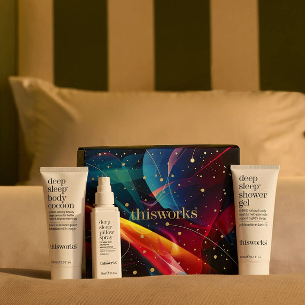 A set of four Thisworks 'Deep Sleep' body care products arranged in front of a colorful cosmic-themed box on a beige surface with a blurred bed background. The set includes a body cocoon, a yellow spray, a shower gel, and the branded box.