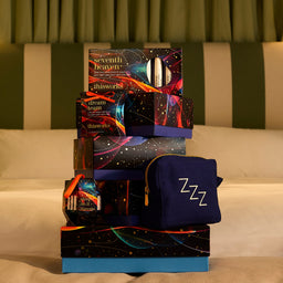 A stacked gift set on a bed featuring multiple cosmetic boxes and a navy blue pouch with 'Zzz' printed on it. The packaging has a cosmic, colorful design with stars and neon lines, and the background shows a green and beige striped headboard.