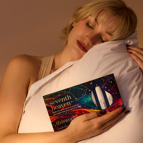 A woman rests her head on a white pillow while holding a colorful product box labeled 'seventh heaven' and 'thisworld' against her chest in warm, dim lighting.