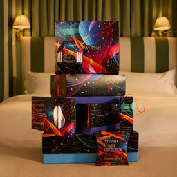 A stack of colorful 'thisworks' and 'dream team' gift boxes arranged on a bed in a bedroom setting. The boxes feature vibrant cosmic and abstract space designs with stars, planets, and neon streaks in blue, pink, orange, and purple.
