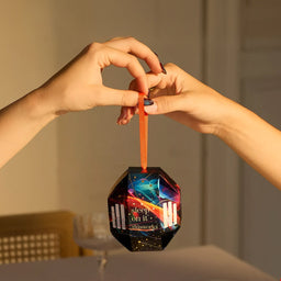 A person's hands hold a faceted black Christmas ornament with a colorful cosmic design and an orange ribbon. The ornament features the text 'sleep on it.