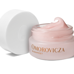 A jar of Omorovicza Budapest cream with its white lid leaning against it on a clean white background. The jar is a soft pale pink, filled with a matching pale pink cream, and features the brand name in gold lettering.