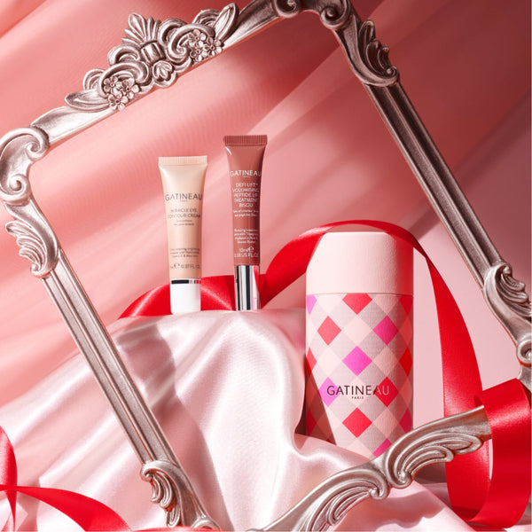 Three GATINEAU cosmetics products arranged on draped pink satin with a red ribbon and an ornate silver mirror frame. The products include a beige tube, a mauve-brown tube, and a pink-and-white argyle cylindrical container.