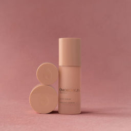 A beige cosmetic bottle with a matching cap stands next to two round beige product pots on a dusty pink background. The bottle features the brand name 'OMORONICA' in black text.