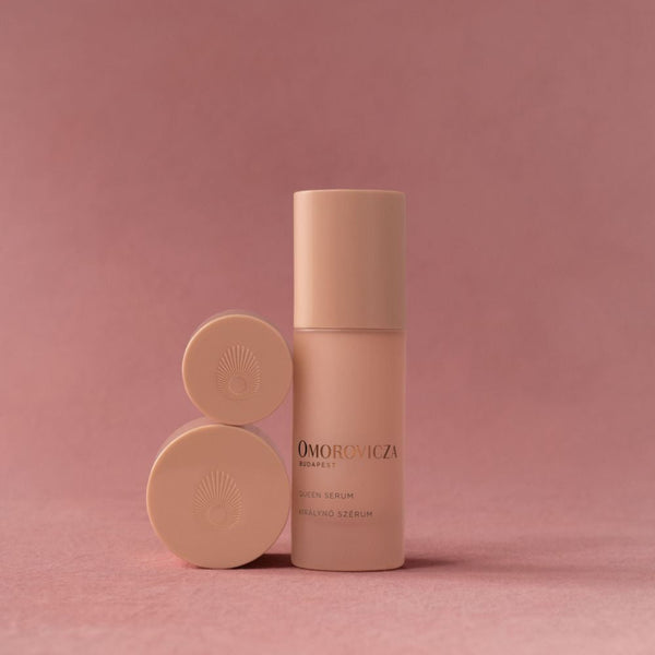 A beige cosmetic bottle with a matching cap stands next to two round beige product pots on a dusty pink background. The bottle features the brand name 'OMORONICA' in black text.