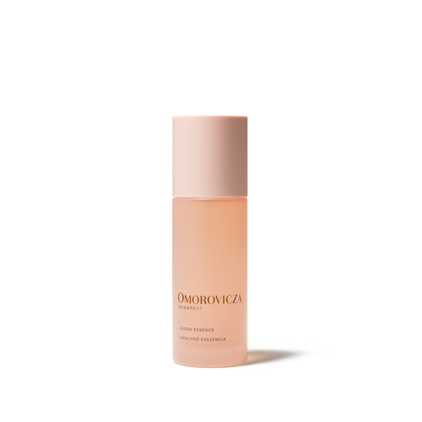 A bottle of Omorovicza Queen Essence Facial Essence in a soft peach-pink shade, featuring a matching pale pink cap, set against a clean white background with a soft shadow.