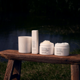 Four white ShimmerBeauty skincare containers arranged on a rustic wooden bench outdoors at night, with grass and a dark background visible. The products include a jar, a candle, a bottle, and two open cream jars.