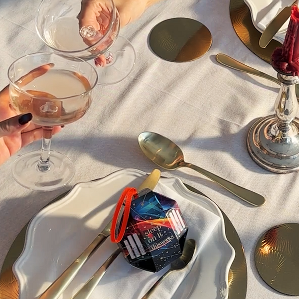 A styled dining table setting with white plates, gold-toned cutlery, gold coasters, and two hands holding stemmed wine glasses. A small packaged item labeled 'SLEEP IN A CASTLE' with a red loop is placed on the tablecloth.