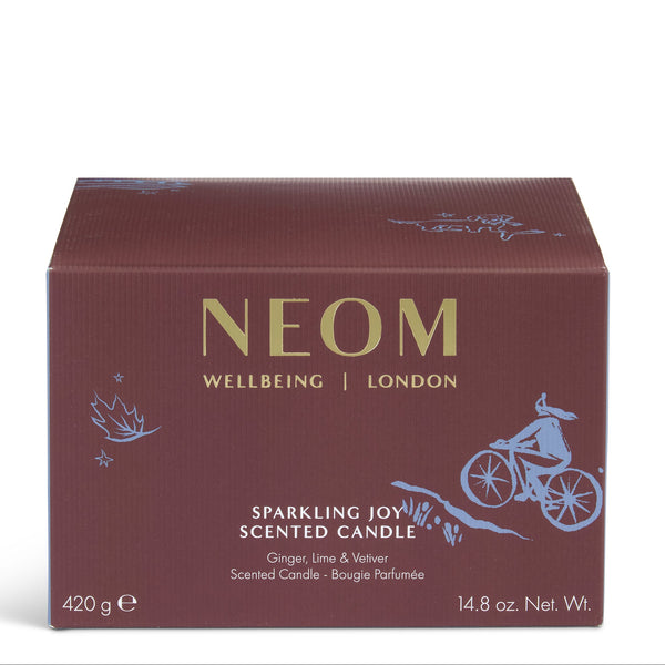 A rectangular NEOM Wellbeing London scented candle box in a deep burgundy-brown color, featuring gold lettering and light blue line-art illustrations of a cyclist and botanical motifs, set against a plain white background.