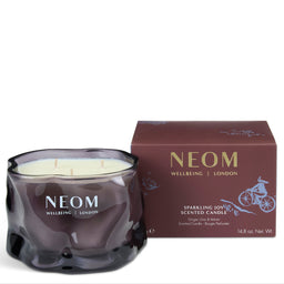 A NEOM Wellbeing London scented candle in a dark, wavy glass jar with cream wax, shown next to its matching dark brown packaging box on a white background.