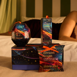 Three beauty products from thisworks arranged on a bed: a faceted glass bottle labeled 'sleep on it', a tall box labeled 'perfect bands', and a box labeled 'dream team'. A person reclines in the softly blurred background.