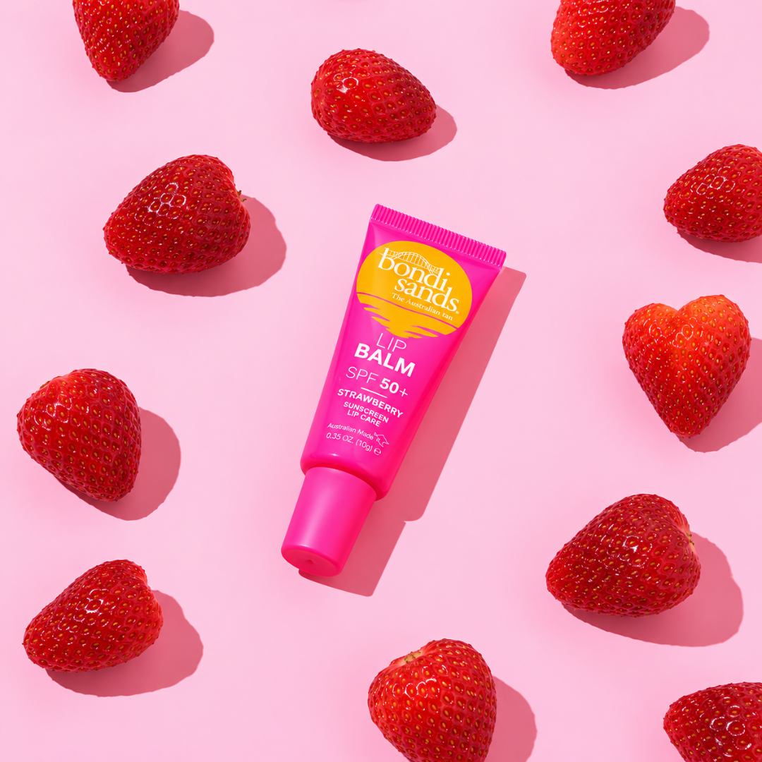 A pink squeeze tube of Bondi Sands Strawberry Lip Balm SPF 50+ lies diagonally on a pink background, surrounded by fresh red strawberries.