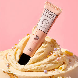 A squeeze tube of Frank | Body Sugar Bark Lip Balm standing upright in a swirl of cream-colored frosting with decorative pearls and gold flakes, against a soft pink background.