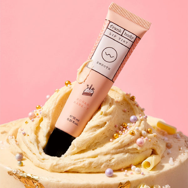 A squeeze tube of Frank | Body Sugar Bark Lip Balm standing upright in a swirl of cream-colored frosting with decorative pearls and gold flakes, against a soft pink background.