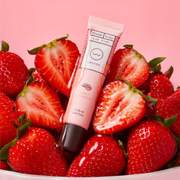 A pink lip tint tube labeled 'fresh body SMOOTH lip tint' standing upright on a pile of fresh sliced strawberries against a solid pink background.