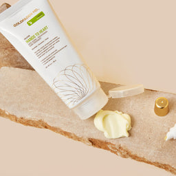 Two Goldencapsules skincare products, Hand & Heart Hand Cream and Lip Therapy Lip Balm, styled on a natural stone slab with a dollop of cream and a gold cap against a beige background.