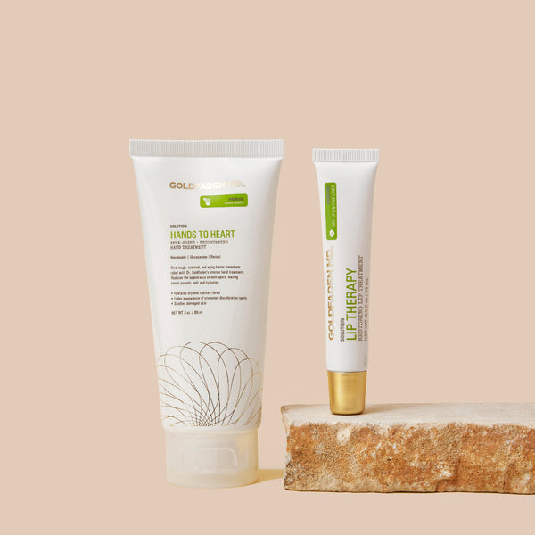 Two white skincare tubes from Dolo Cosmetics, including Hands to Heart Hand Cream and Lip Therapy, displayed on a textured stone slab against a solid beige background.