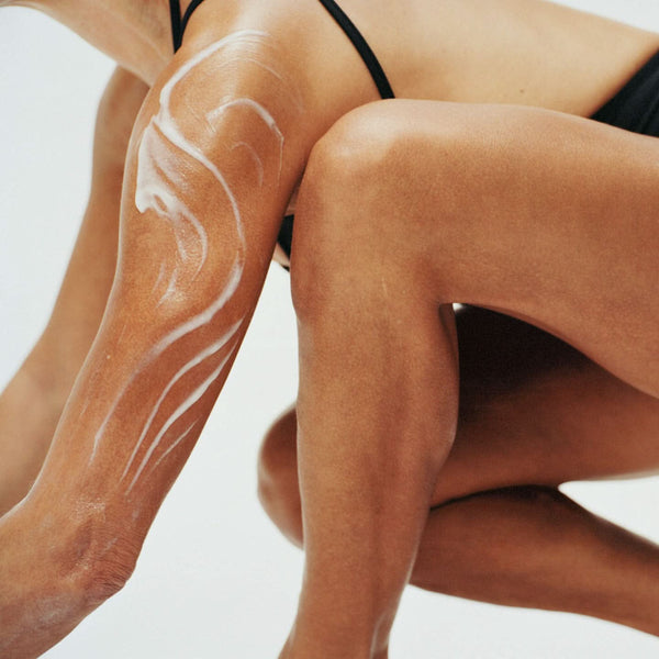 Close-up of a person's legs and torso wearing black bikini bottoms, with white lotion or sunscreen swirled onto the skin against a clean white background.