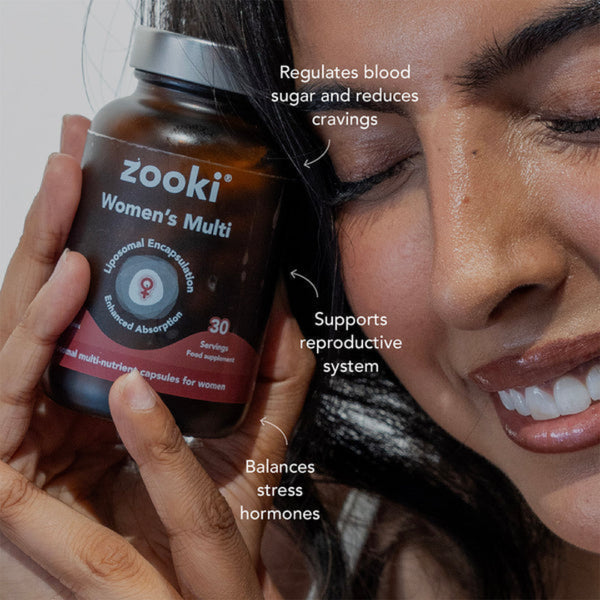 A close-up lifestyle shot of a woman holding a bottle of zooki Women's Multi supplements. The bottle is dark brown with a silver cap and features a label with a red band at the bottom.