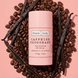 A pink Frank Body caffeine deodorant stick sits in the center, surrounded by scattered coffee beans and vanilla beans on a soft pink background.