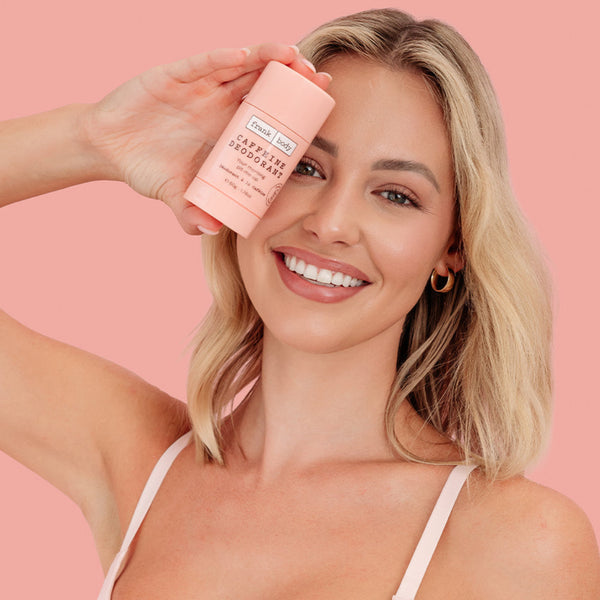 A smiling blonde woman in a pale pink top holds a pink deodorant stick to her face against a solid pink background. The product label reads 'The Deodorant Company' and 'Caramel Apple Deodorant'.
