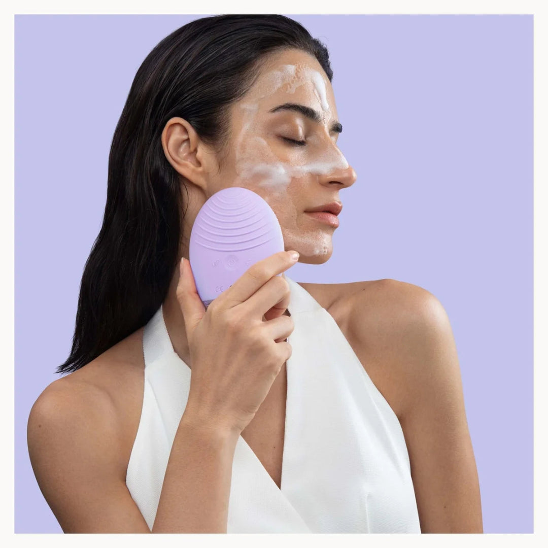 A woman with white face cream on her cheek holds a lavender purple silicone facial cleansing brush against her face, set against a solid light purple background.